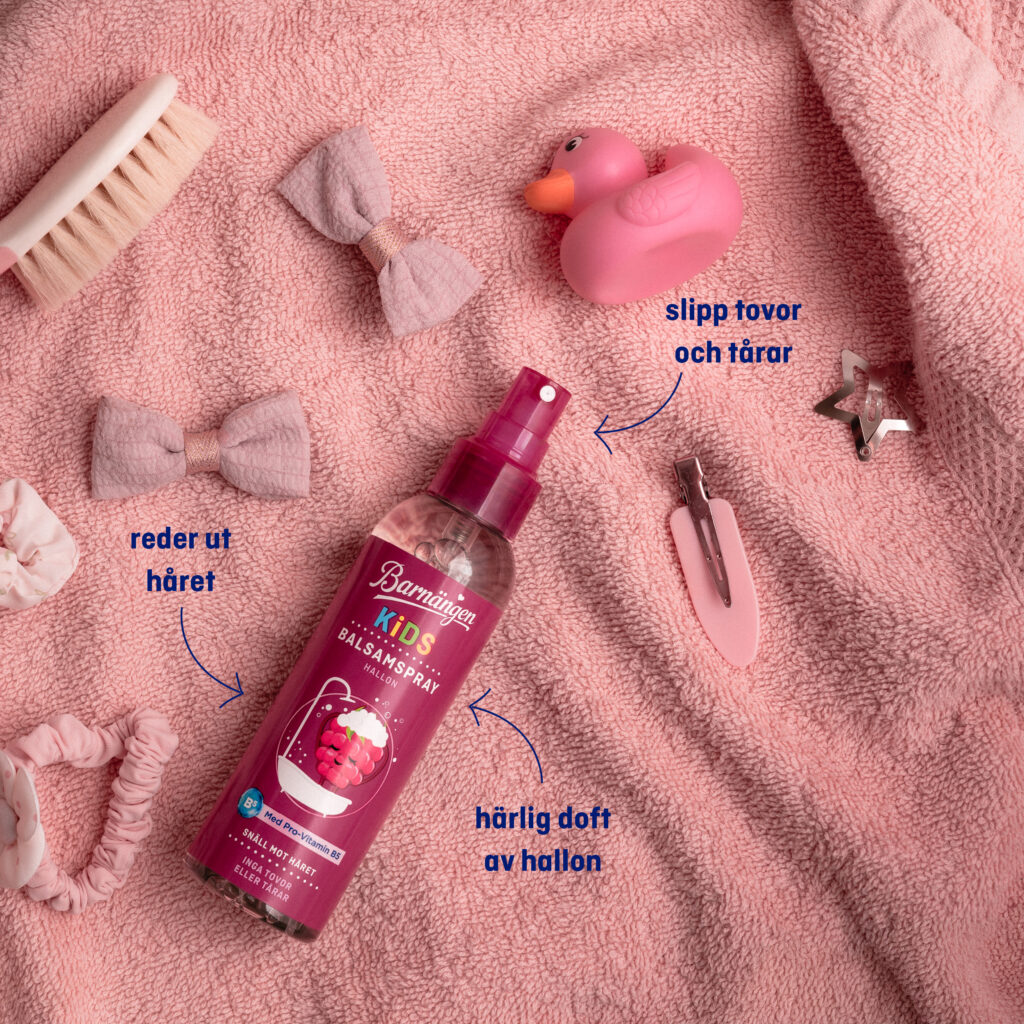 Barnängen balsamspray för barn med härlig doft av hallon ligger på en rosa handduk, omgiven av en badanka, lite hårspännen och en hårborste för barn